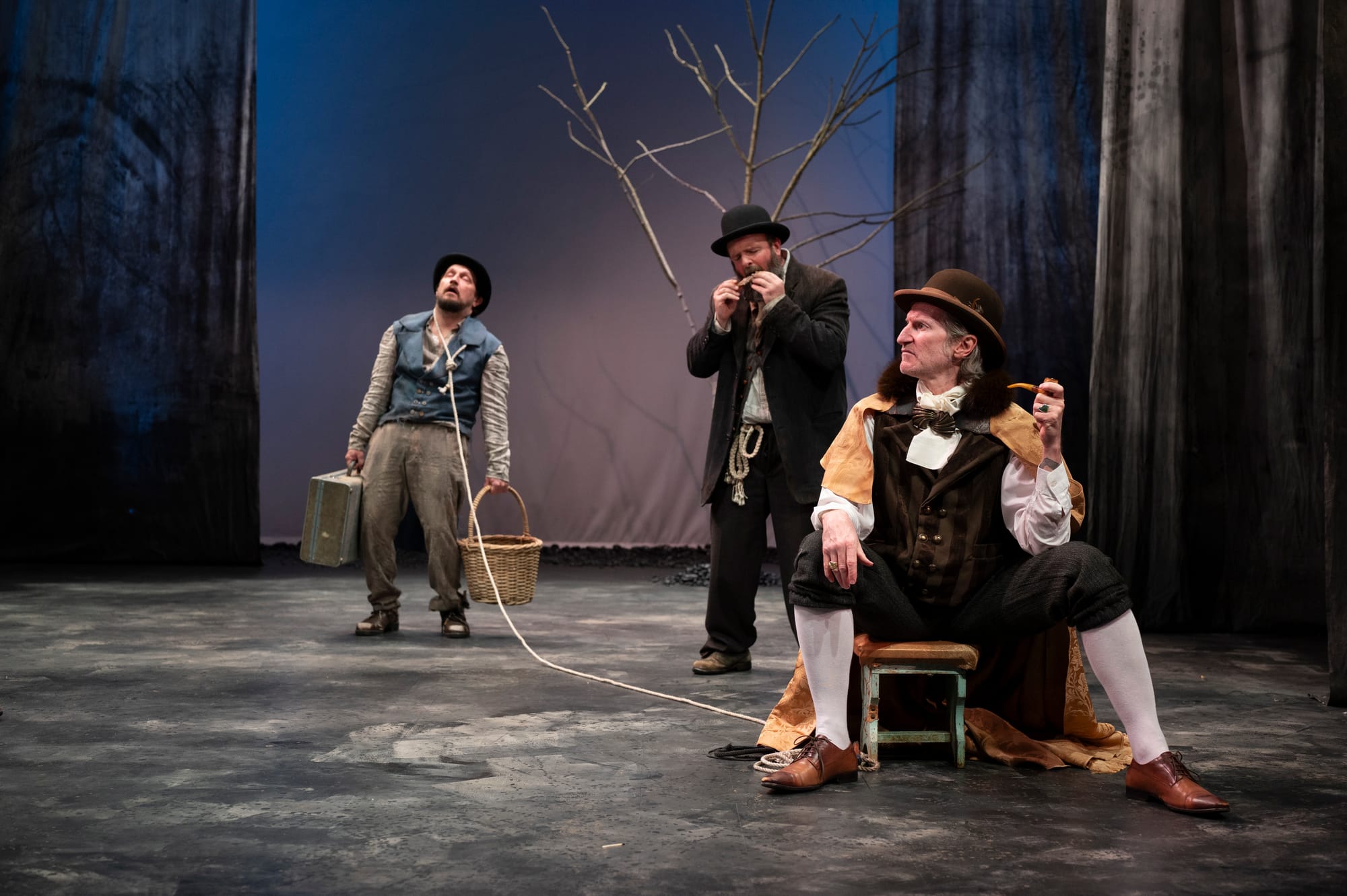 Lucky stands exhausted at left holding a suitcase and basket, tethered by a long white rope to Pozzo, who sits at right in a fur-trimmed cape and bowler hat. Estragon stands between them beneath the bare tree.