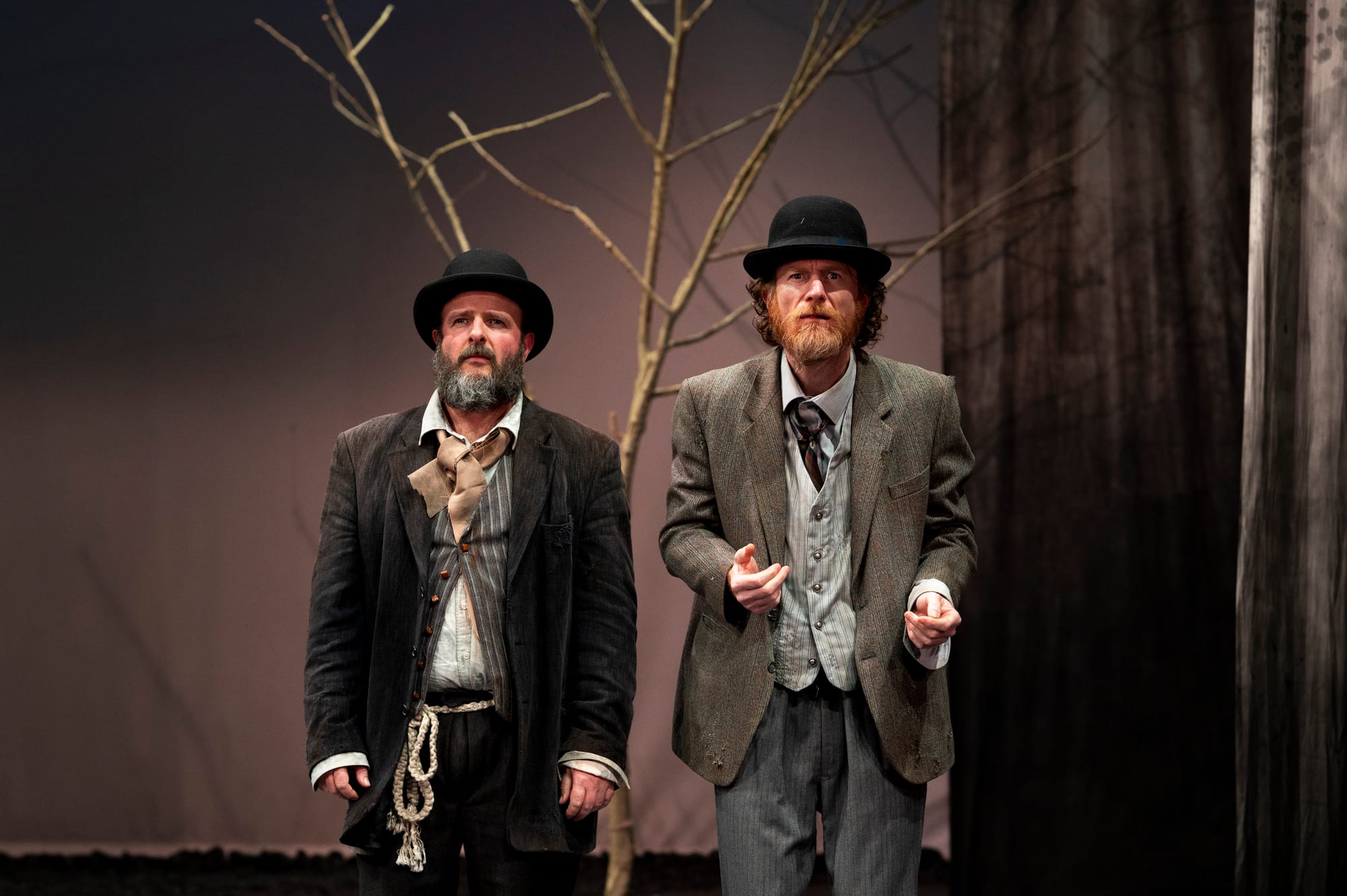 Estragon and Vladimir stand together beneath the bare tree. Estragon, in a dark rumpled suit and bowler hat, looks wearily ahead while Vladimir leans forward beside him with an intent expression.
