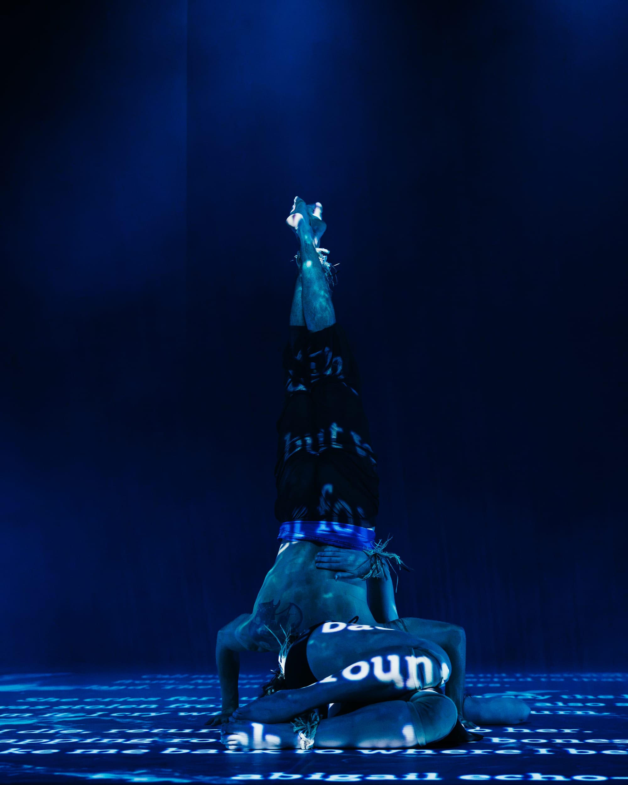 One dancer holds a vertical inverted balance above another, who is folded low on the floor. Blue light and projected words cover their bodies and the surrounding stage.