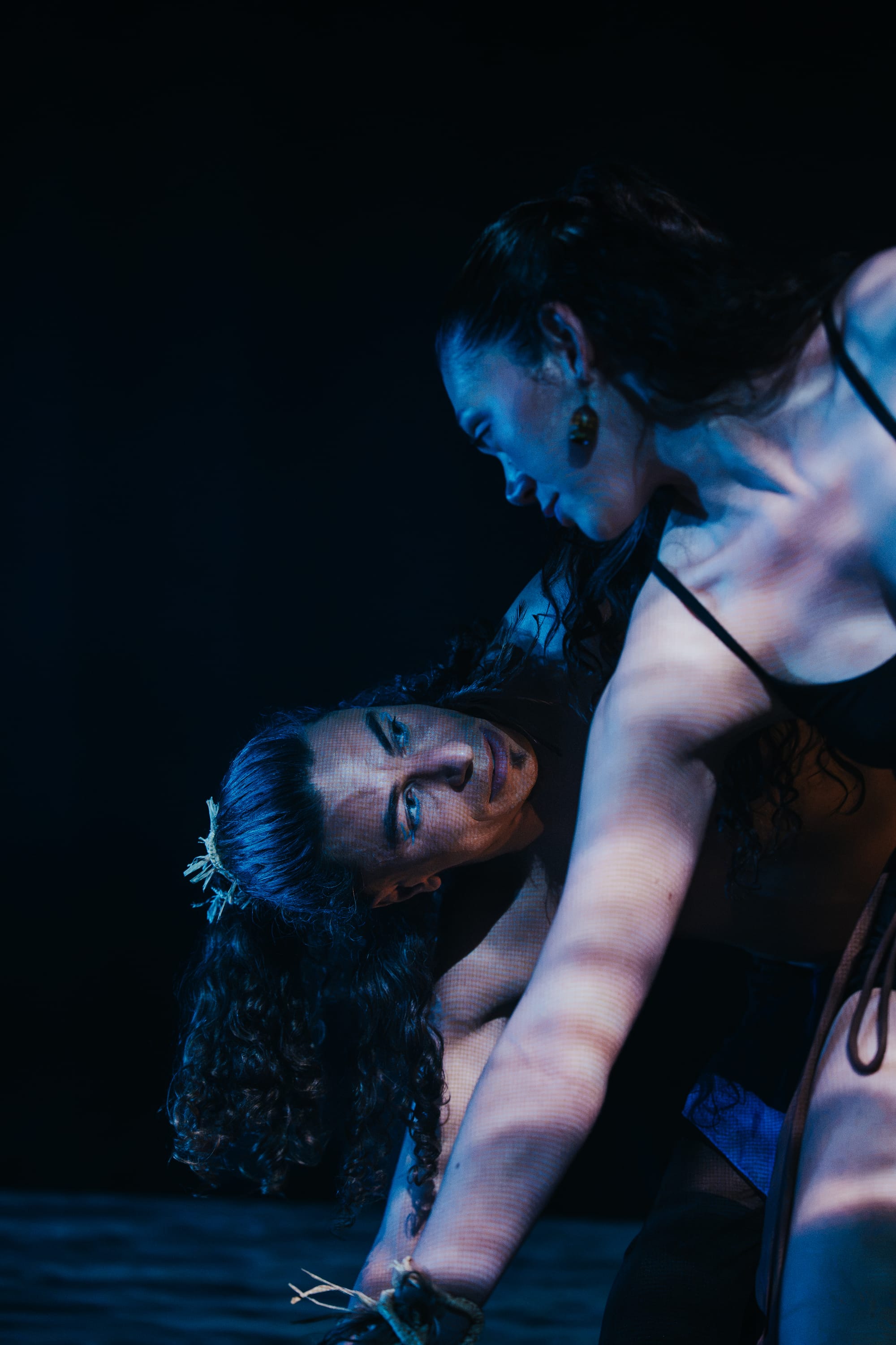 Close-up of two dancers leaning across one another under blue light. One looks down intently while the other, partly reclined beneath them, smiles upward.