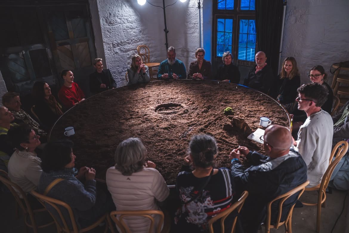 Luke Casserly and audience members sit closely around a large circular table covered entirely in bog soil during Distillation.