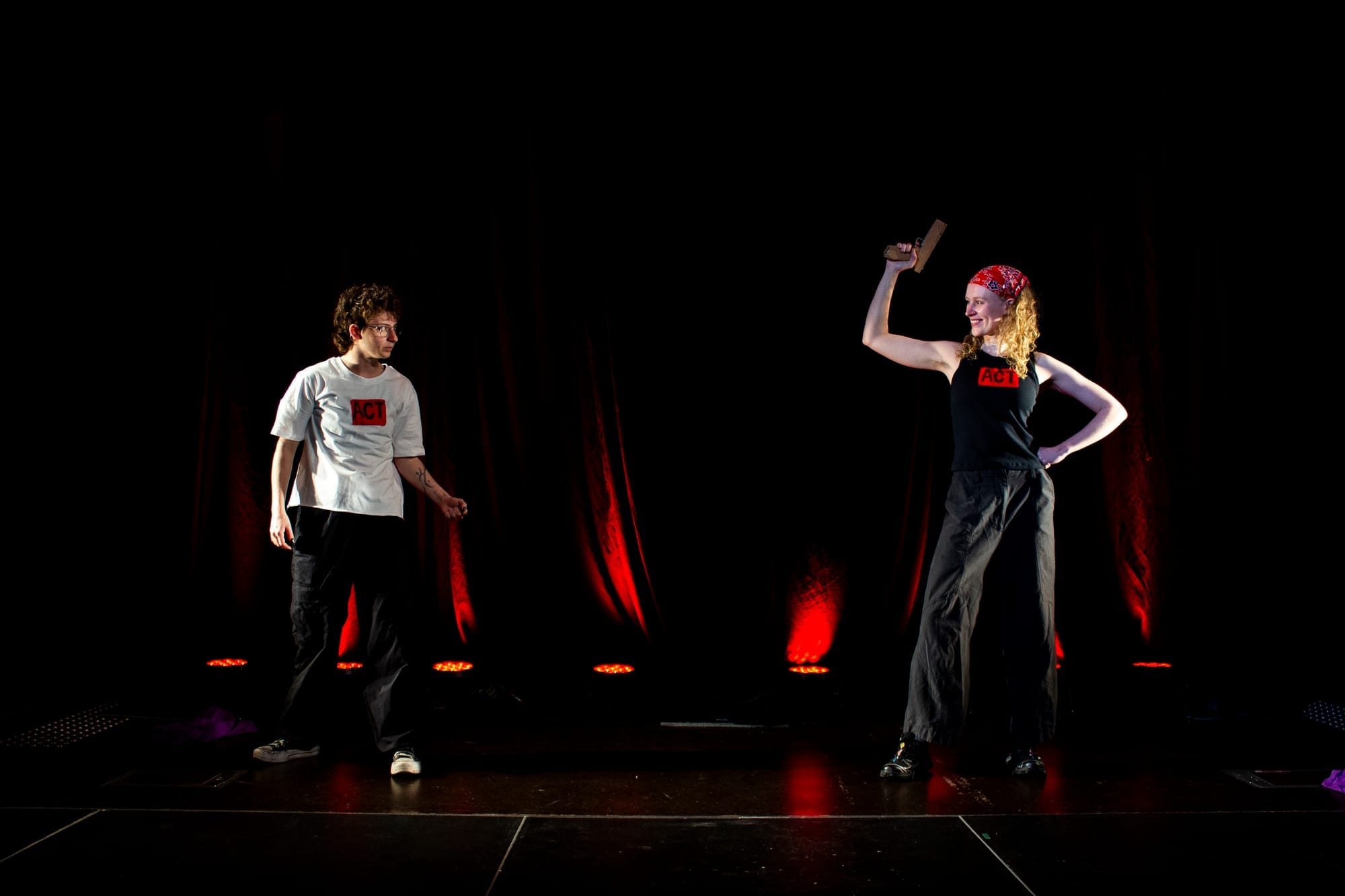 Two ACT members face each other on a dark stage lit from behind in red; one holds a gun aloft.