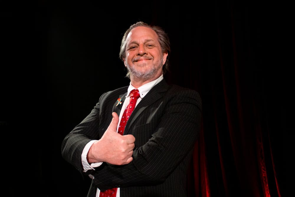 Scott Gooding as Arts Minister Richard Head stands in a dark suit and red tie, arms folded and giving a broad thumbs-up to the audience.