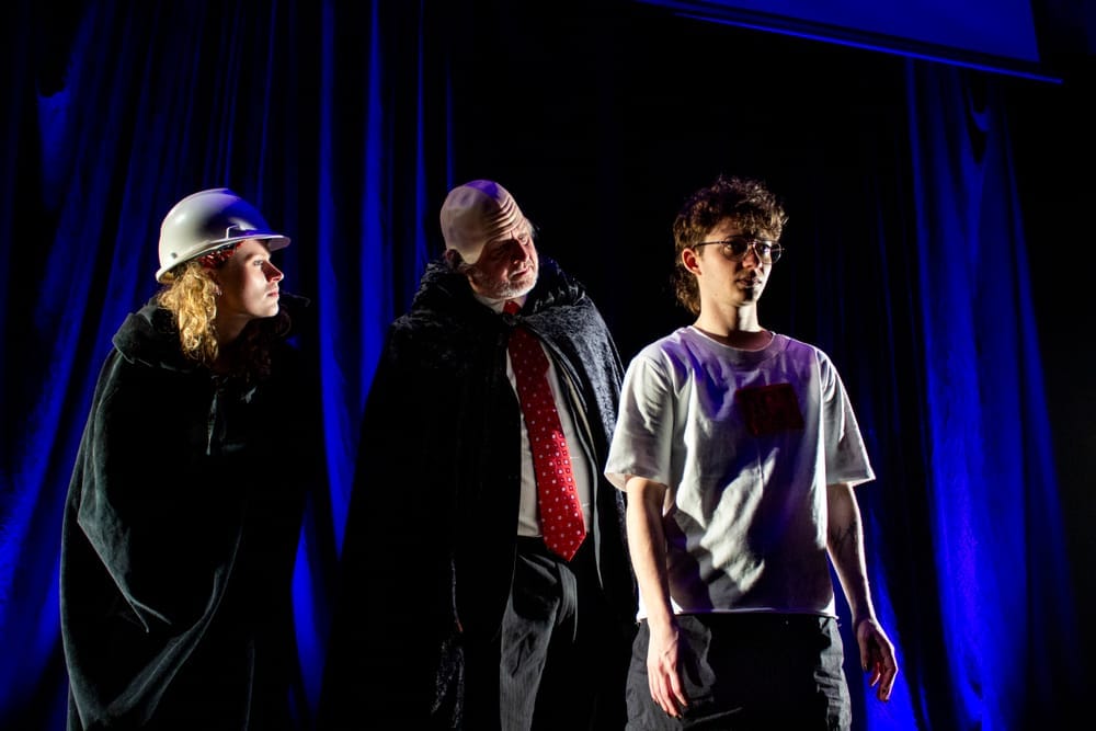 Two performers appear as the Weird Sisters over Chase's shoulder, under blue stage lighting, wearing an assortment of cloaks and headwear.