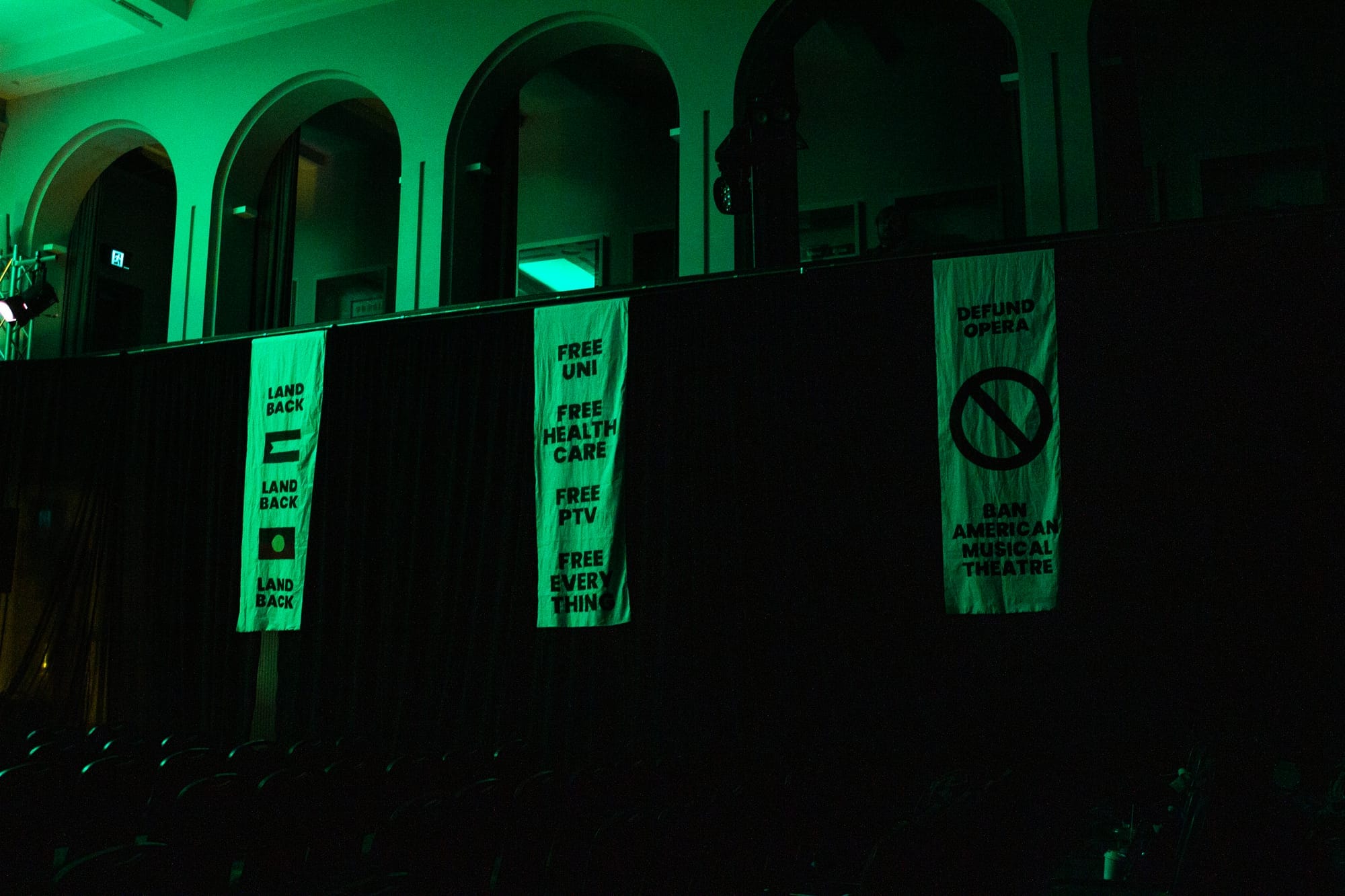 Protest banners hang from the Trades Hall balcony, including “Land Back”, “Free Uni”, “Free Health Care”, “Free PTV”, “Free Everything”, “Defund Opera” and “Ban American Musical Theatre”.