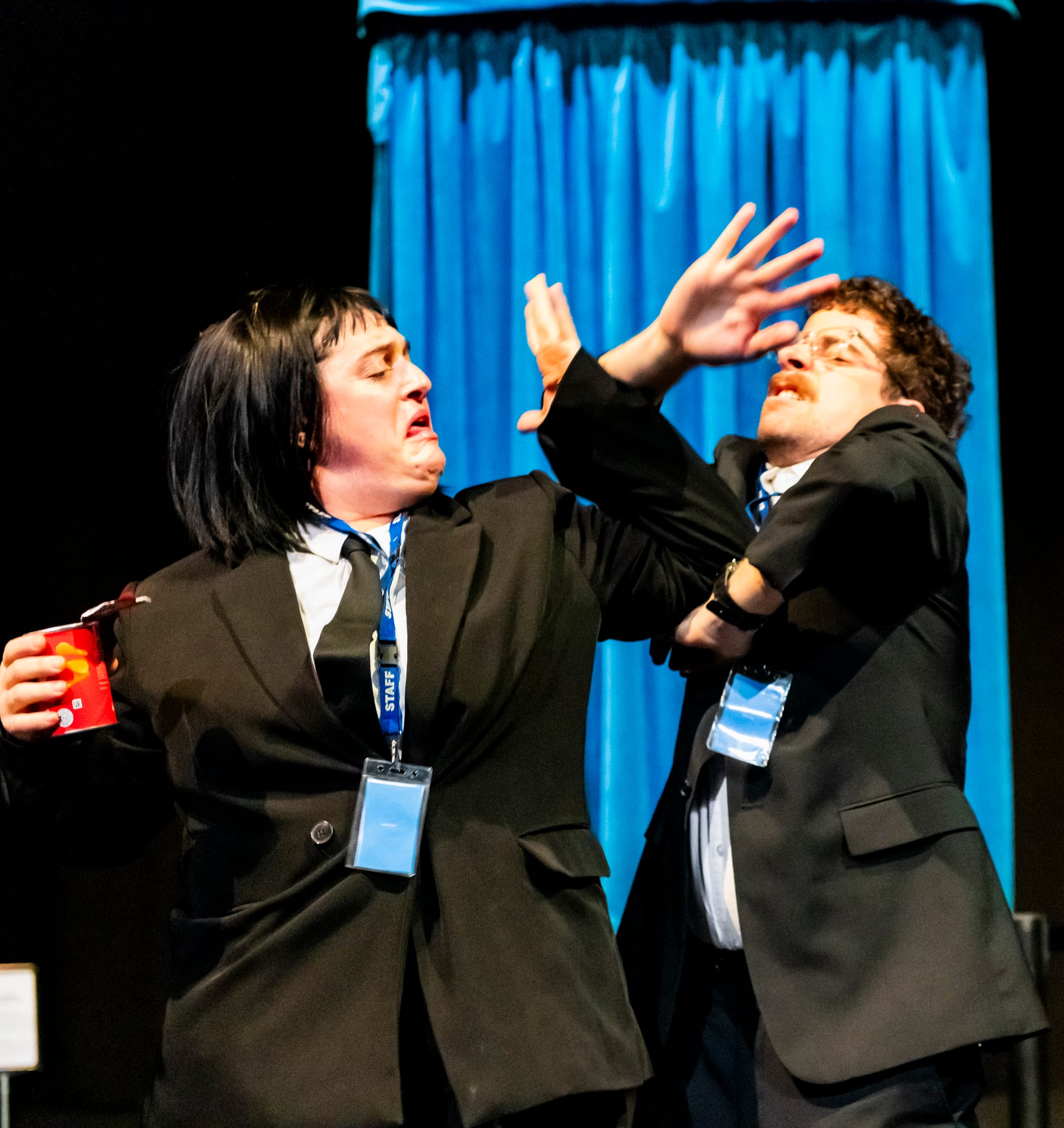 Emilie Bloom and Tim Sneddon tussle in front of a bright blue curtain, Bloom holding a red cup while Sneddon recoils with his arms raised.
