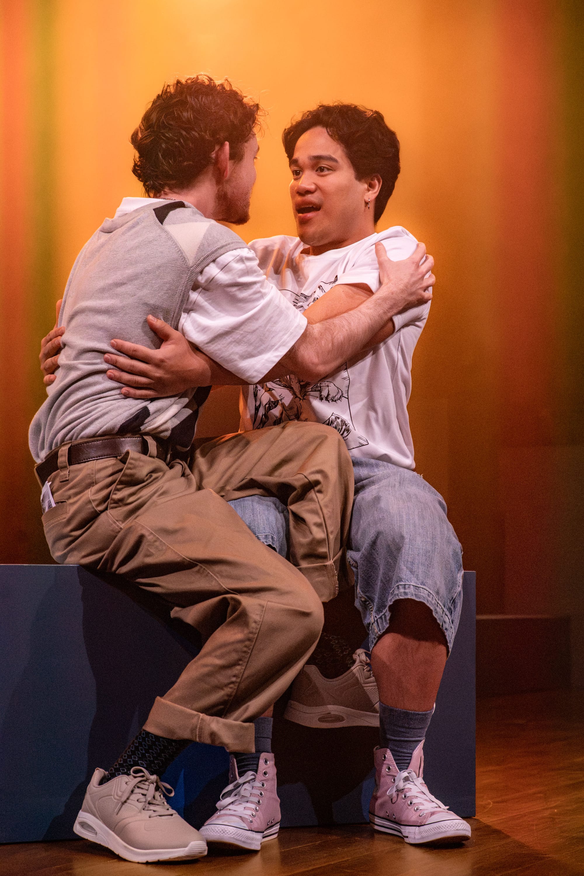 Lawrence Hawkins and Ken Paolo sit pressed closely together on a blue block, arms around each other, against a vivid orange-lit background.