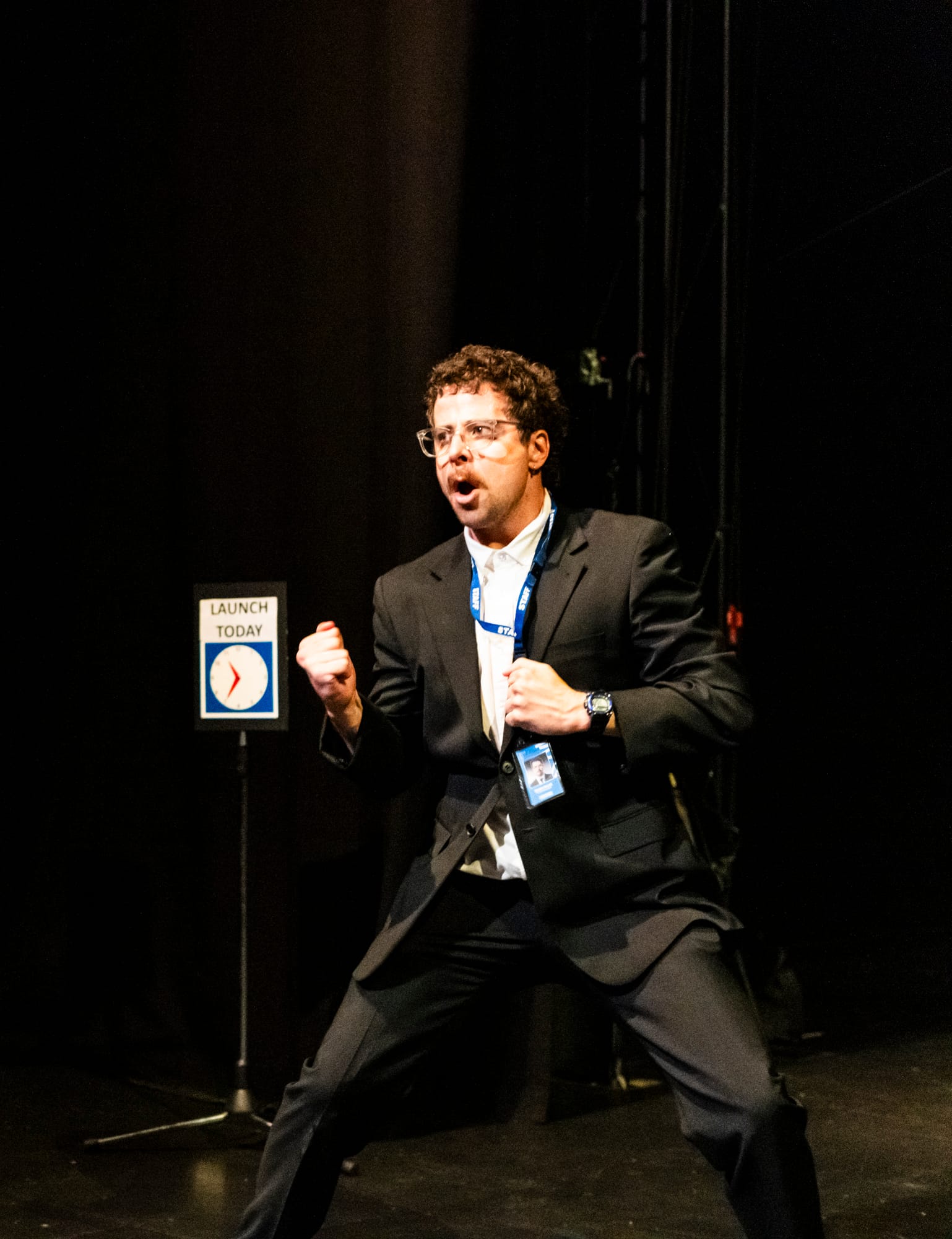 Tim Sneddon, dressed as a gallery attendant in a black suit and staff lanyard, stands with legs apart and one fist clenched, his mouth open in an exaggerated expression of triumph.