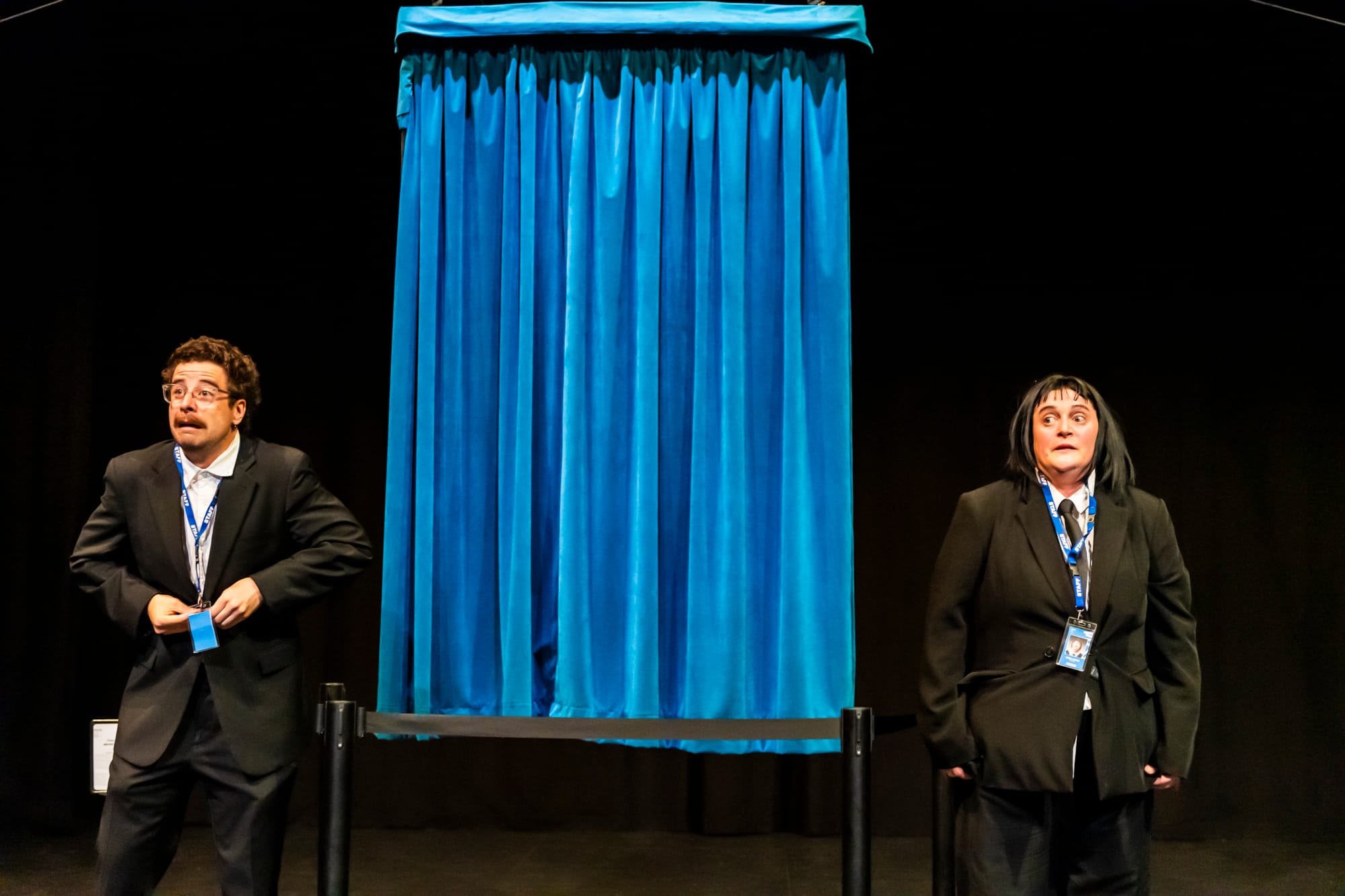 Tim Sneddon and Emilie Bloom stand stiffly on either side of a tall blue curtain concealing the portrait, both wearing black suits and blue staff lanyards with anxious expressions.