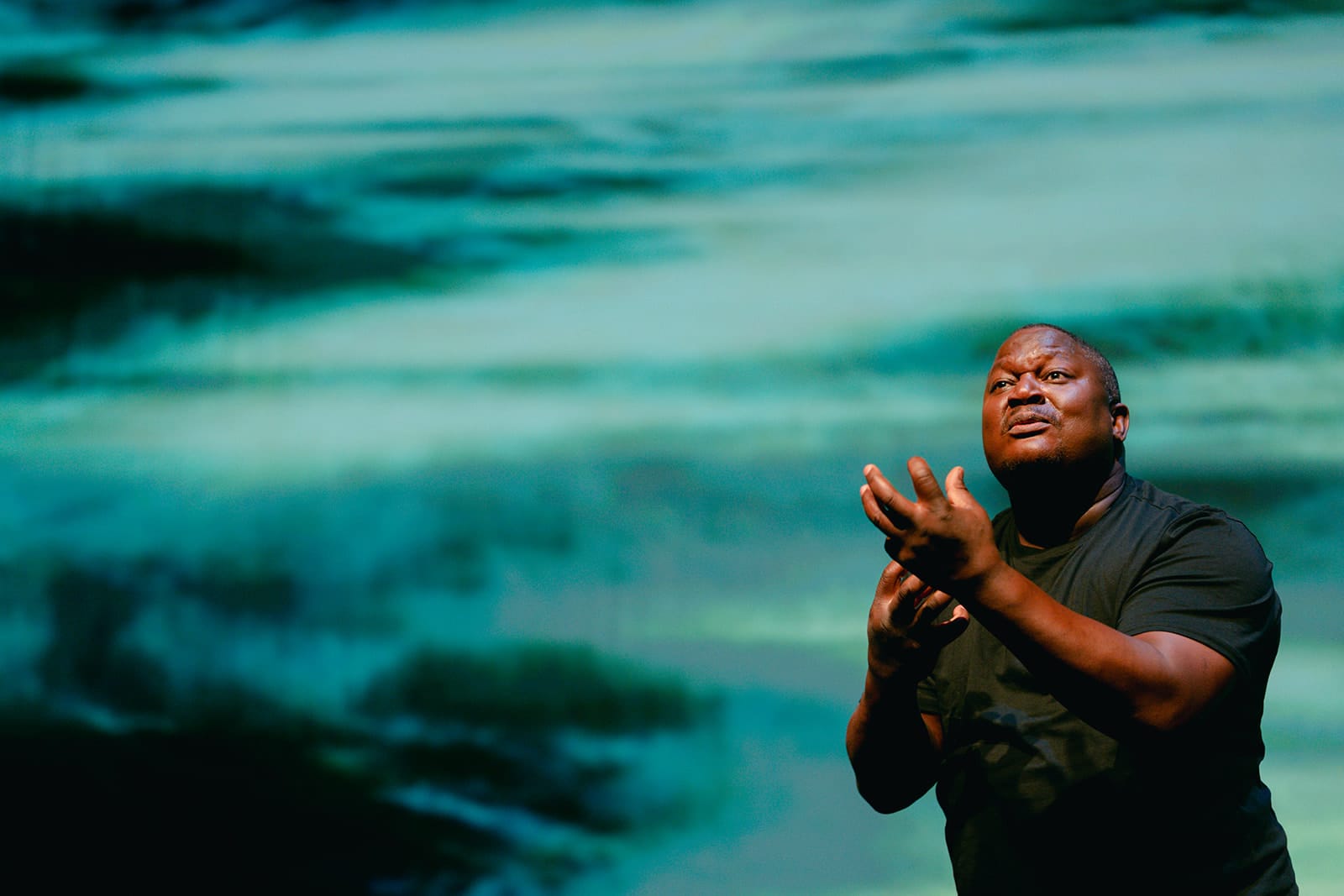 Walter Kadiki signs expressively onstage, his hands raised and face turned upwards, against a large projection of swirling turquoise ocean water.
