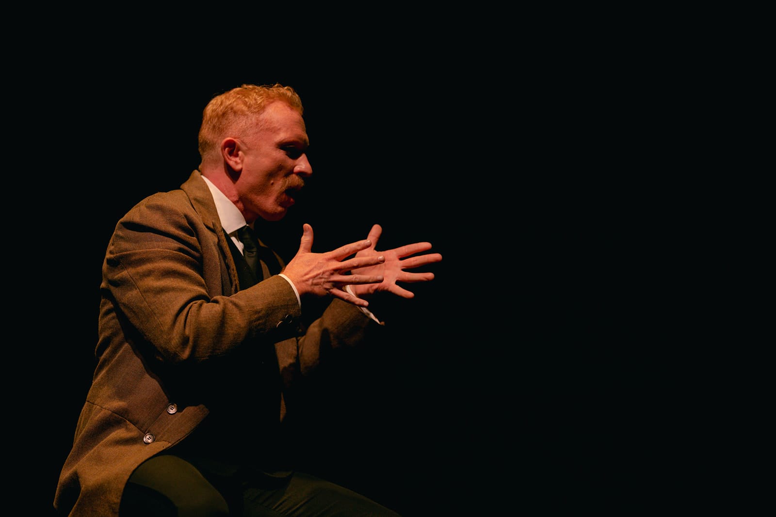 A performer with short light-coloured hair and a large moustache sits onstage in a brown period suit, signing emphatically with both hands against a black background.