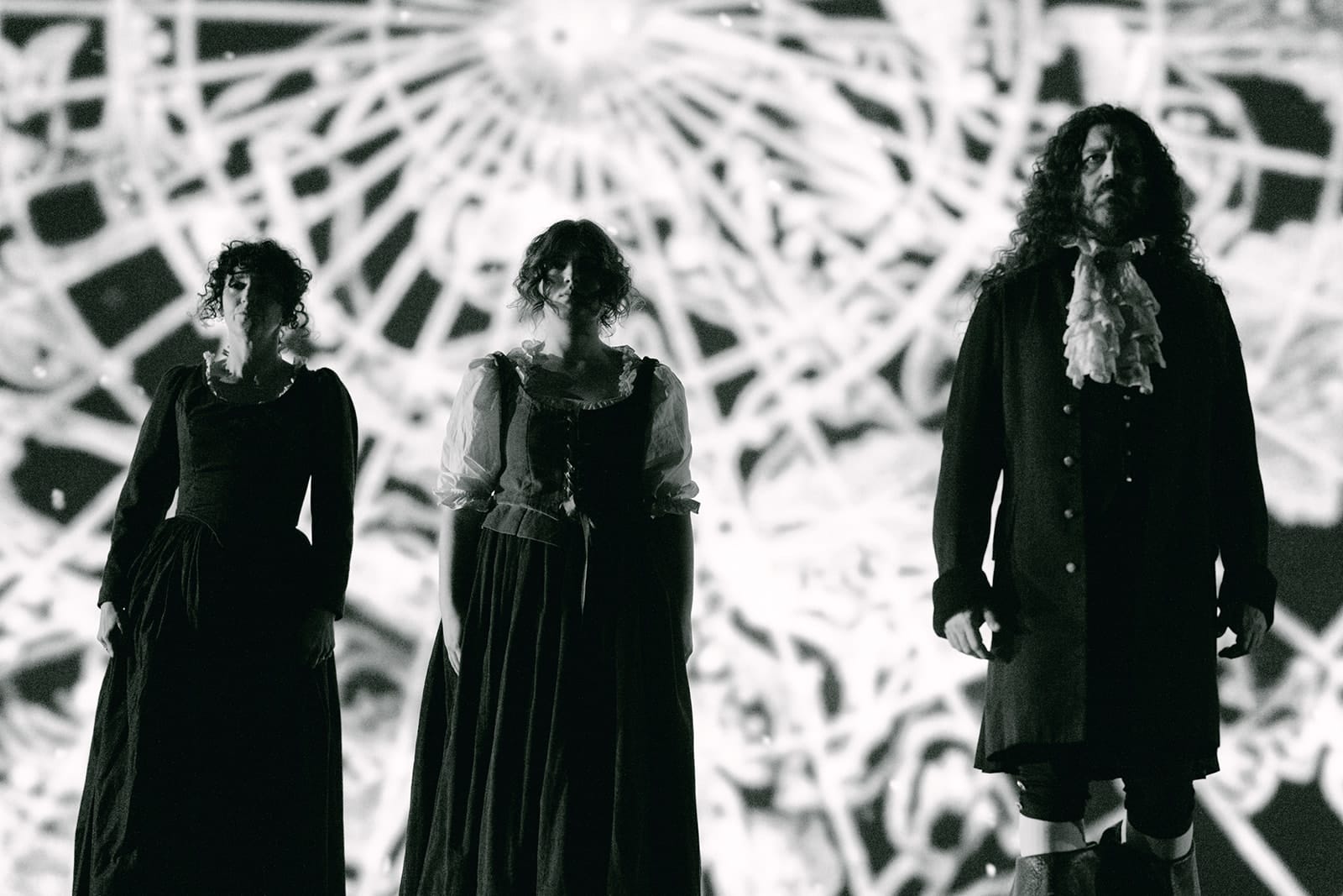 Three performers stand in eighteenth-century-style costumes before a large black-and-white projection of an intricate circular diagram. Two wear long dresses and the third a dark coat with a white cravat.