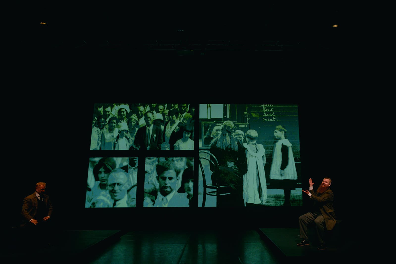 A darkened stage with two performers positioned on opposite sides of a large projection showing a collage of historical black-and-white photographs, including portraits and classroom scenes.