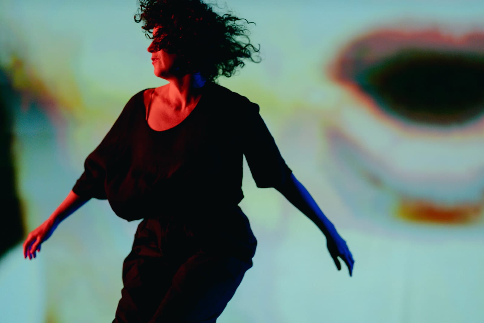 Anna Seymour dances with her arms extended and curly hair flying across her face, illuminated by red and blue light. A large, blurred projection of a mouth fills the background.