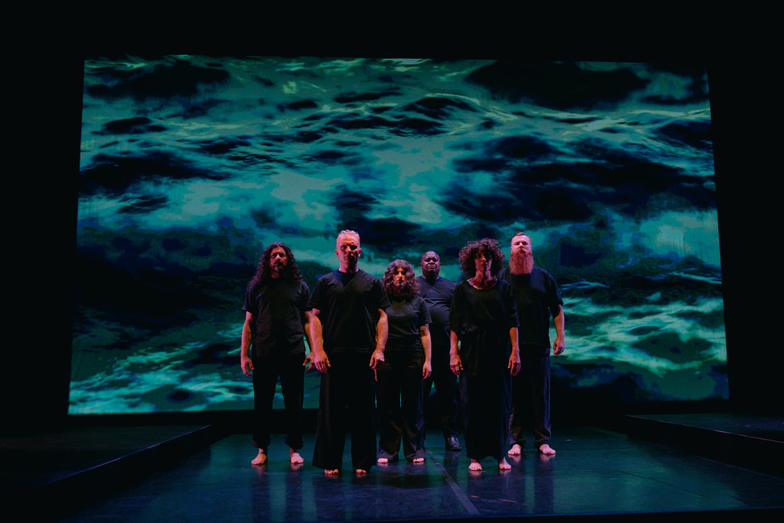 Six performers dressed in black stand together on a dark stage, facing the audience. Behind them, a large projection shows turbulent turquoise ocean waves.