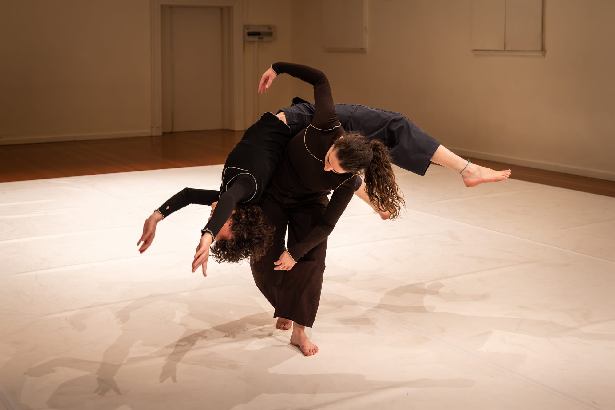 Two barefoot dancers in dark clothing share a balance, one draped sideways across the other’s bent back, with arms and a leg extended.