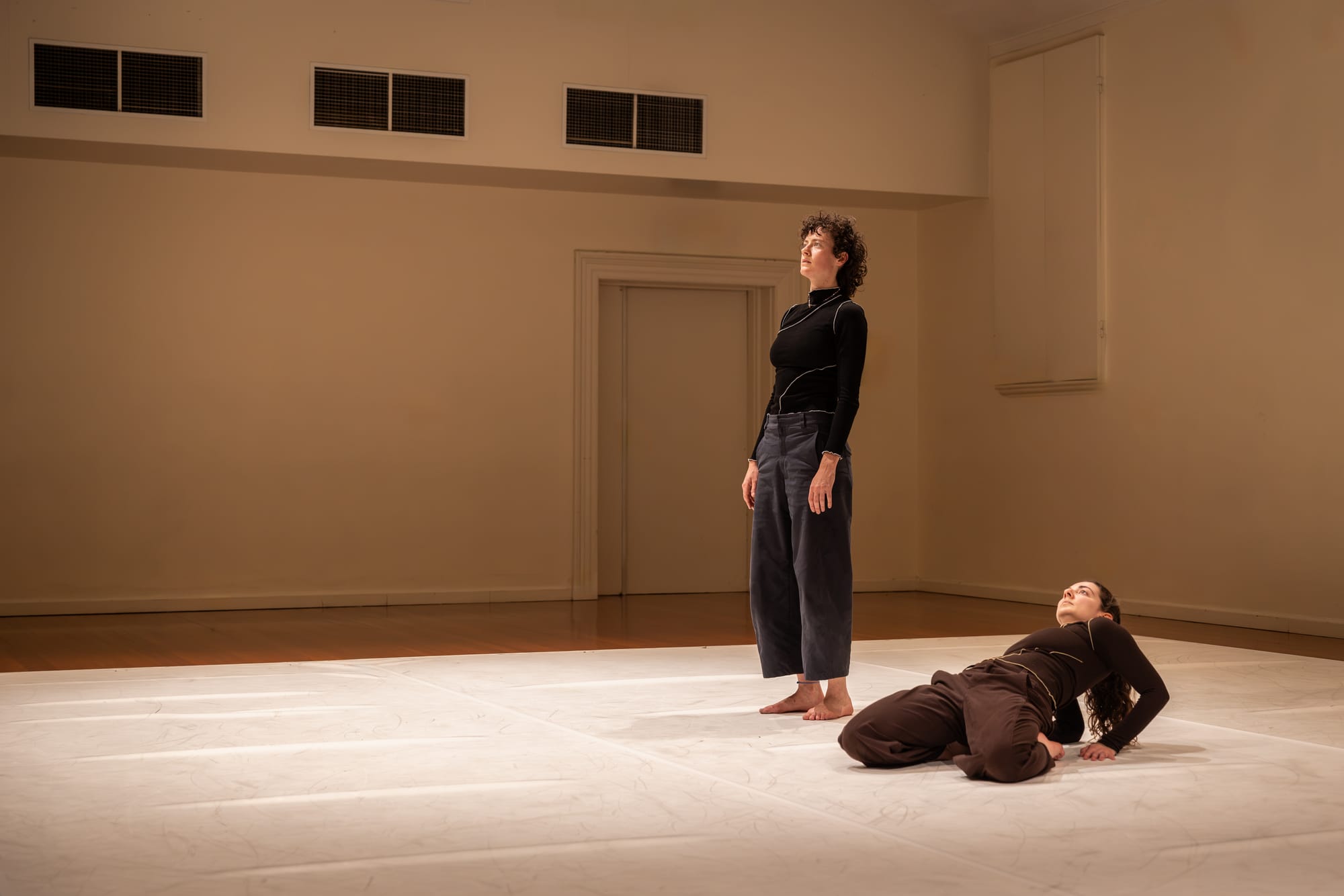 One dancer stands upright with arms at her sides while another reclines beside her, propped on her hands, in a warmly lit studio.