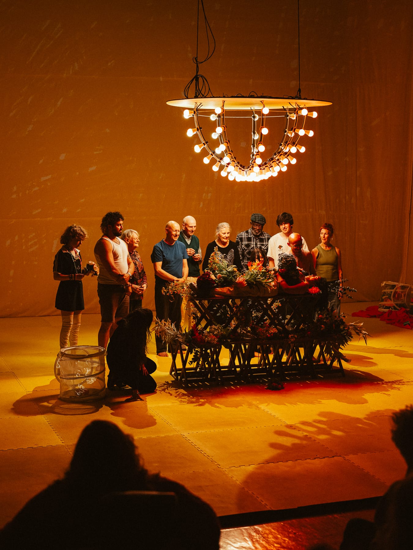 Performers of different ages gather around a flower-covered platform beneath a large illuminated chandelier, surrounded by the yellow stage design of Prime Time.