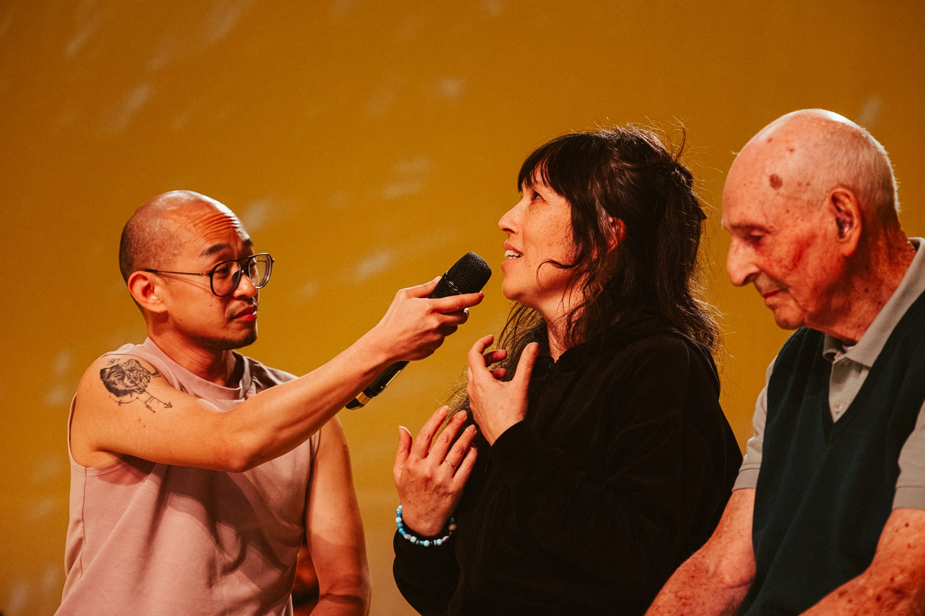 Three performers in Prime Time: one holds a microphone towards a woman speaking animatedly, while an older man sits beside her against a warm yellow backdrop.