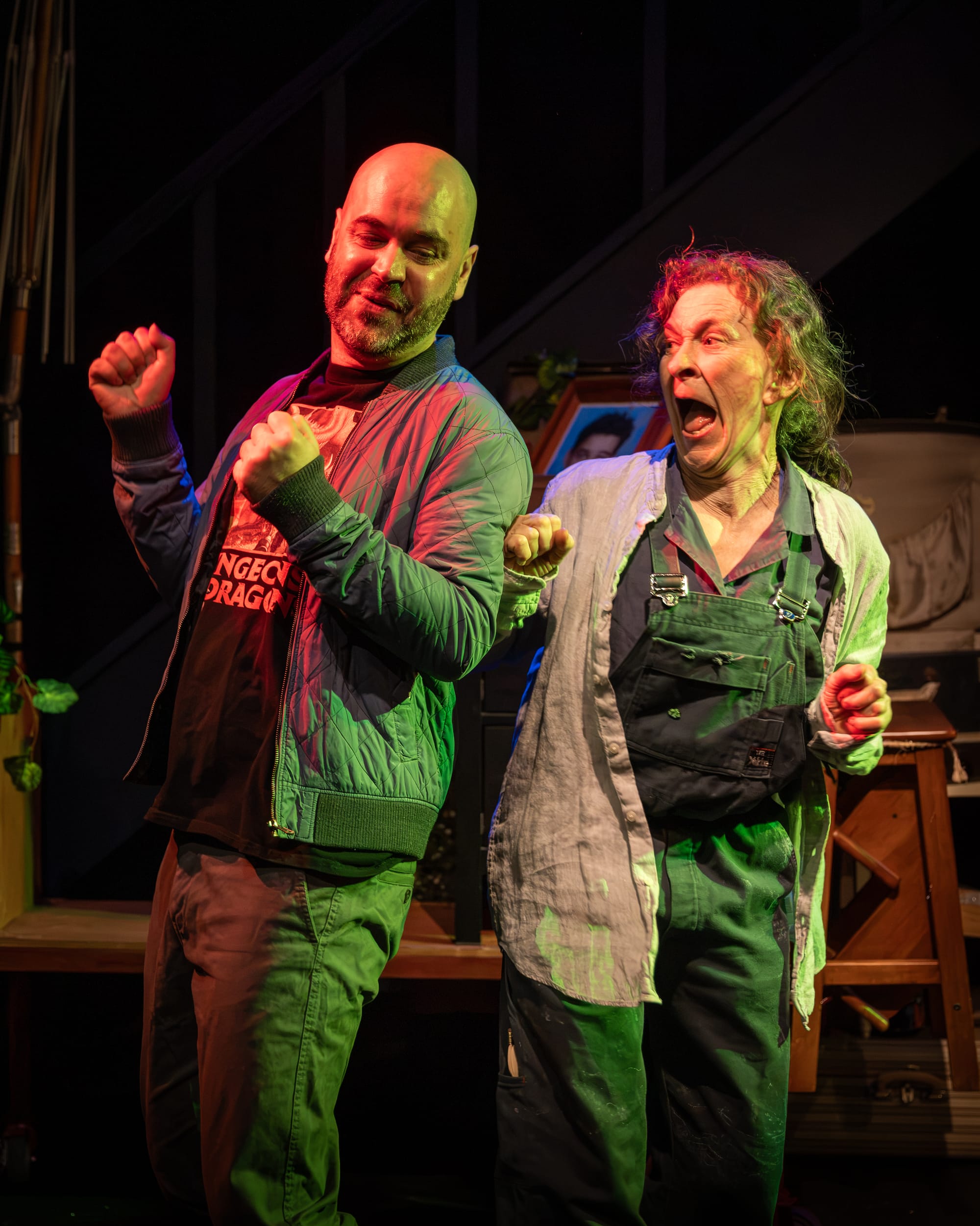Alex Duncan and Felicity Steele dance side by side, their arms raised and faces animated, under vivid red and green stage lighting.