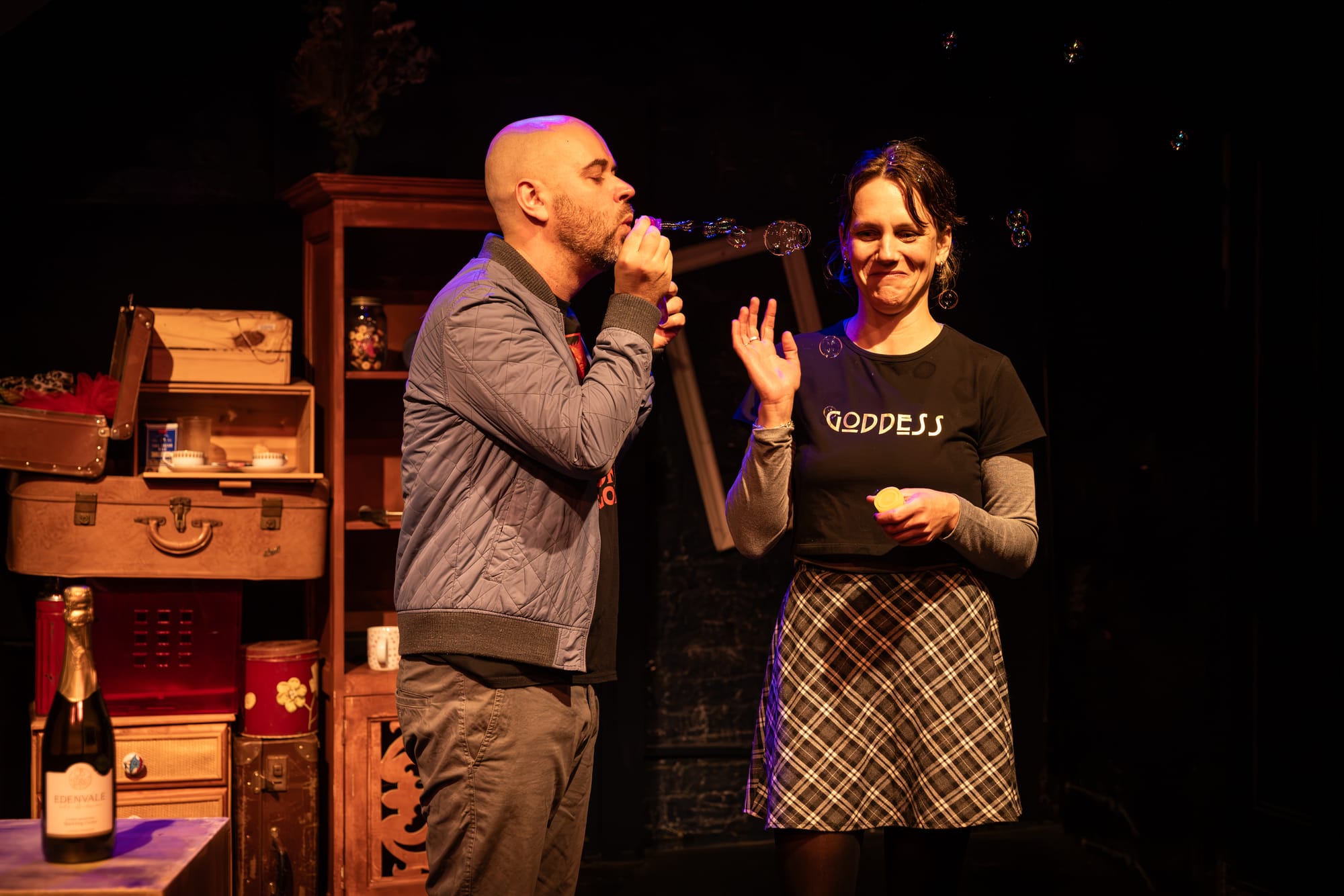 Alex Duncan as Phillip blows soap bubbles towards Michelle Myers as Yvonne, who smiles and raises one hand. Suitcases and mismatched furniture fill the stage behind them.