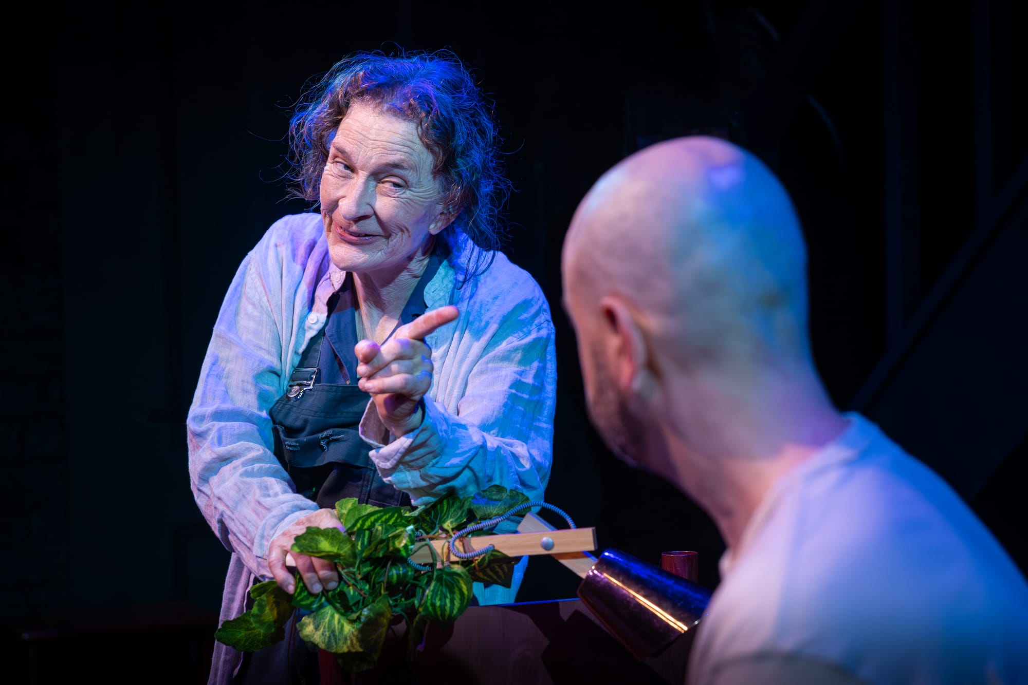 Felicity Steele as Toto smiles mischievously and points towards Phillip, played by Alex Duncan, while holding a leafy green plant under blue stage lighting.
