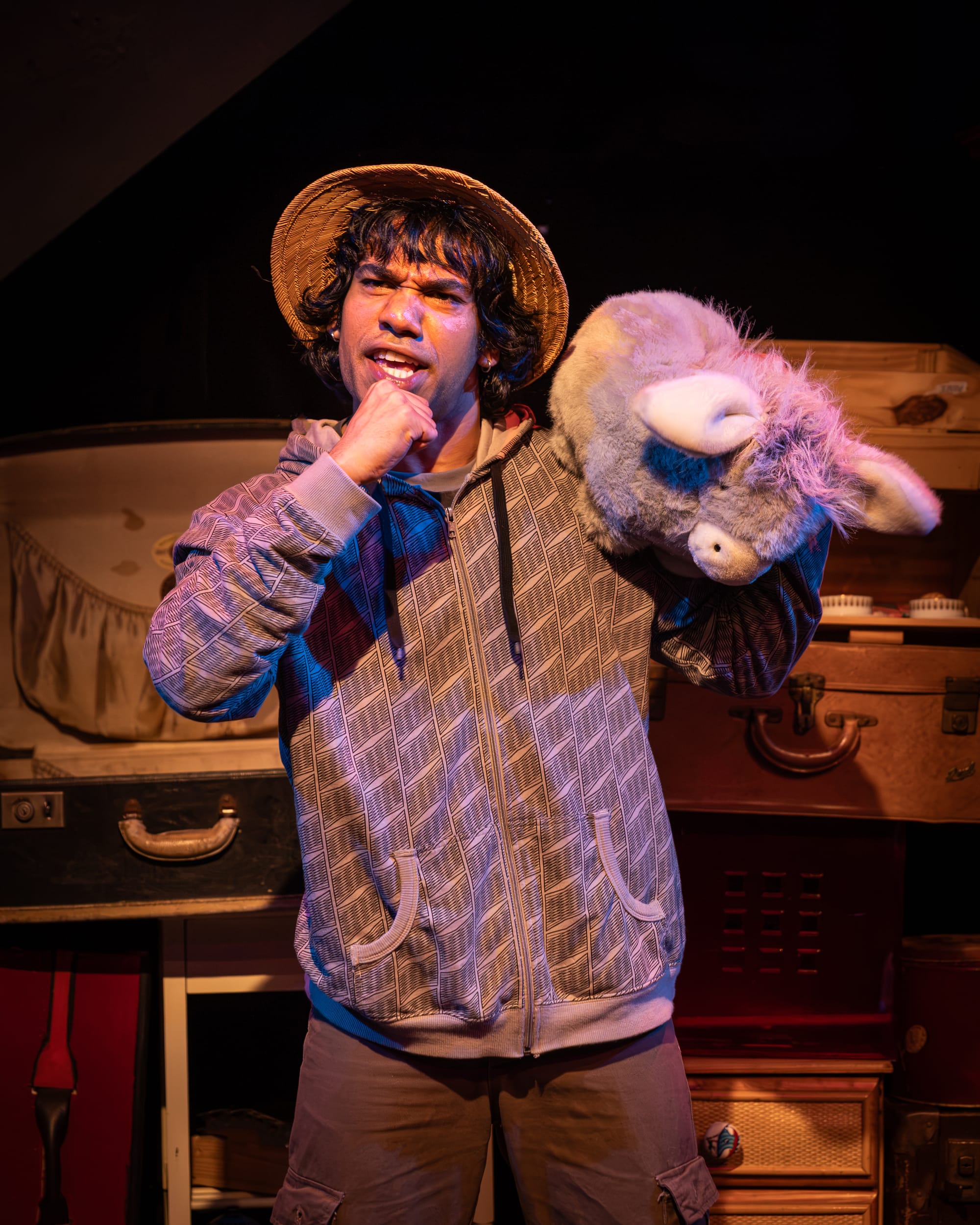 Indra Liyanaarachchi as Solomon wears a straw hat and patterned hoodie, holding a large plush animal over one shoulder and singing with an imaginary microphone.