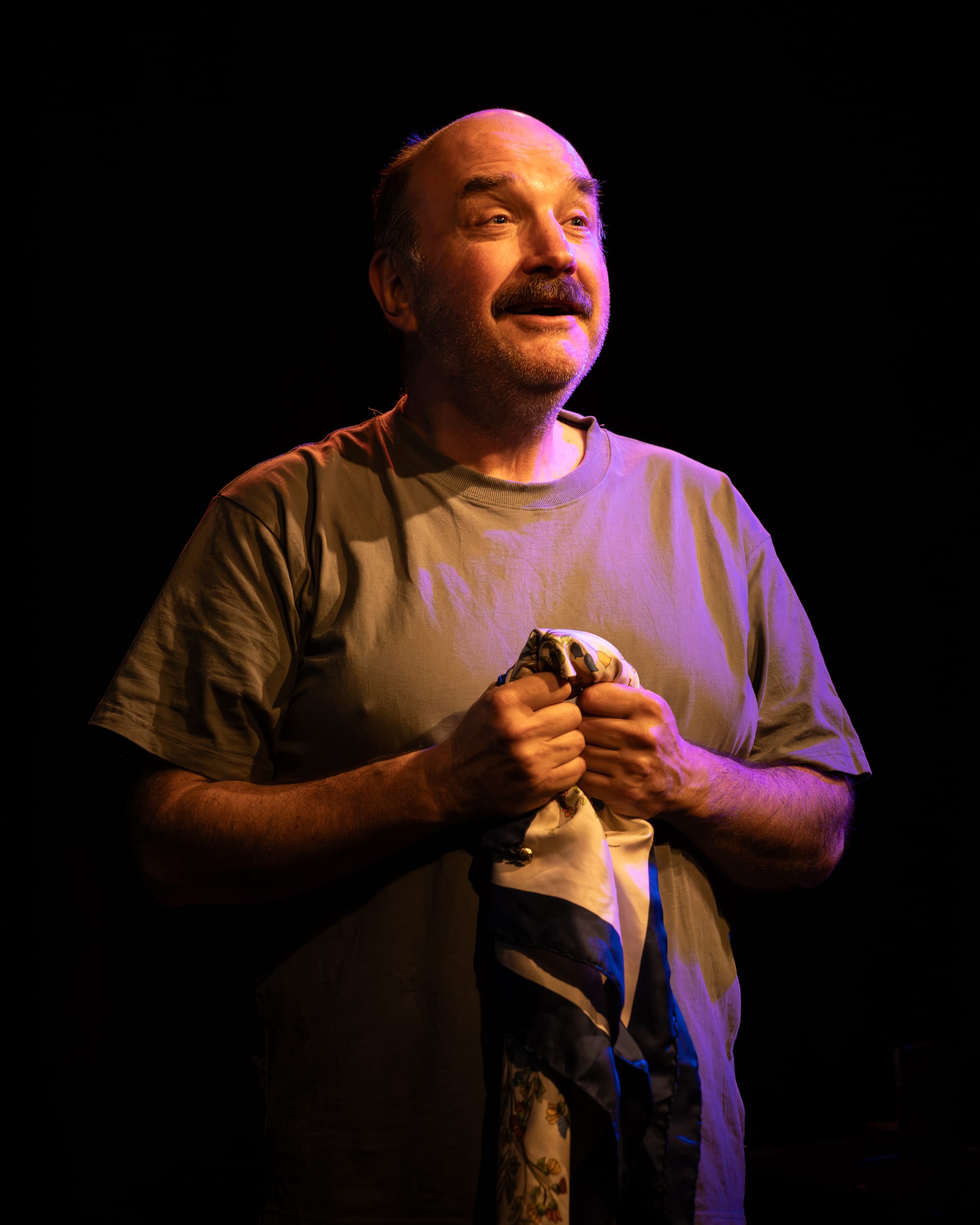 Ben Grant holds a scarf close to his chest during Wrong Way Grow Back at La Mama HQ, warmly lit against a dark stage.
