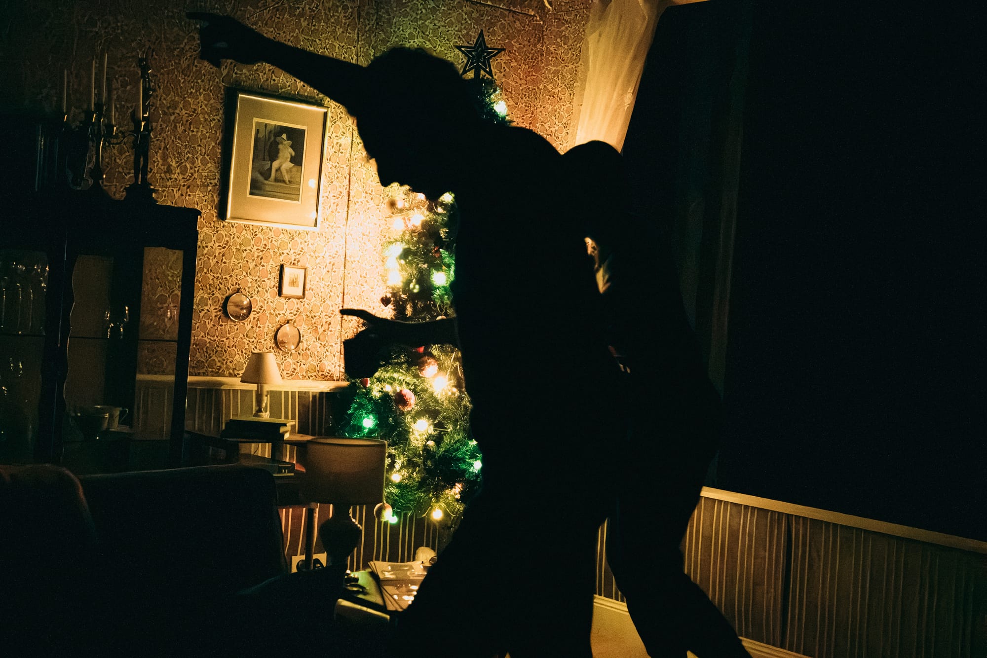 Silhouetted figures dance in a dimly lit living room. A decorated Christmas tree glows behind them, surrounded by vintage furniture and patterned wallpaper.