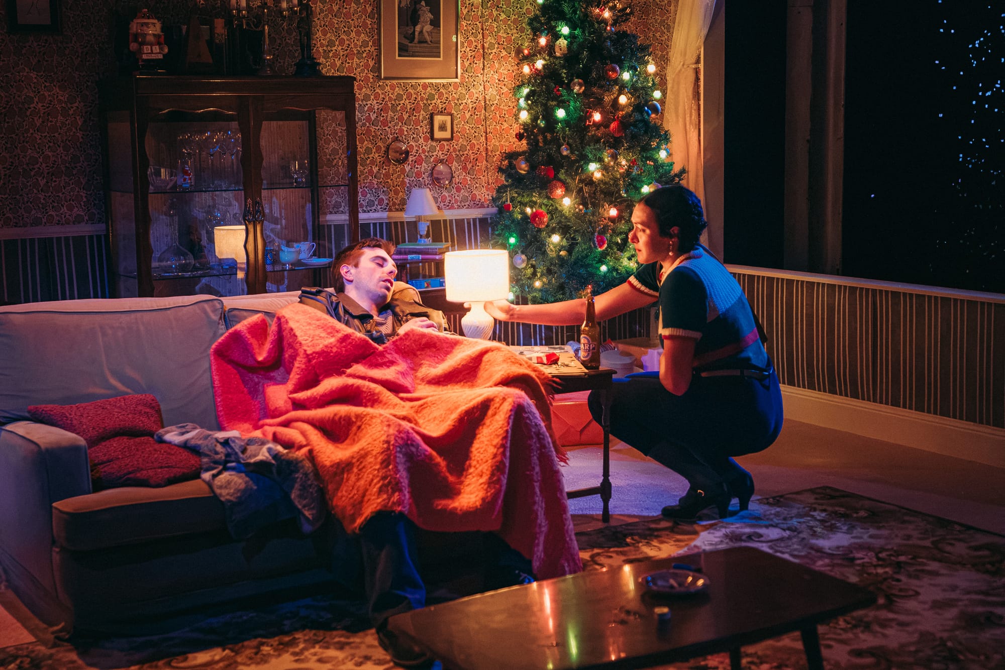 Damon Baudin lies asleep on a sofa beneath a red blanket while Ella Ferris crouches beside him, reaching towards a glowing lamp. A decorated Christmas tree stands behind them in the warmly lit living room.