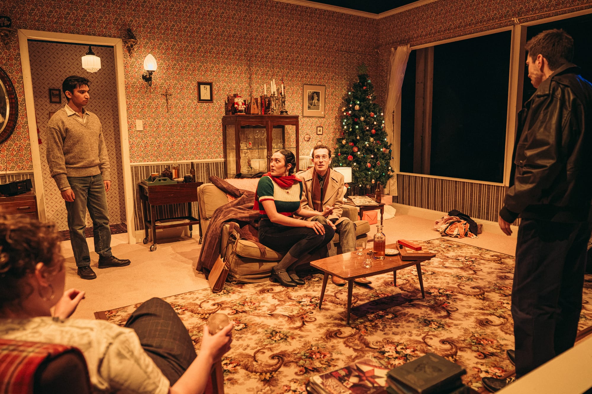 Five performers occupy a richly furnished living room with floral wallpaper, a patterned rug and a Christmas tree. Two sit together on a sofa while others stand or sit around them, their attention directed across the room.