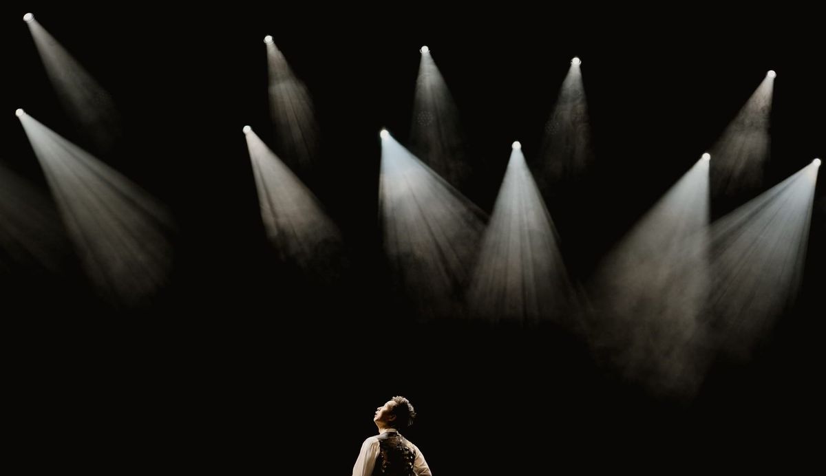 Performer standing beneath a series of white stage spotlights against a dark background.