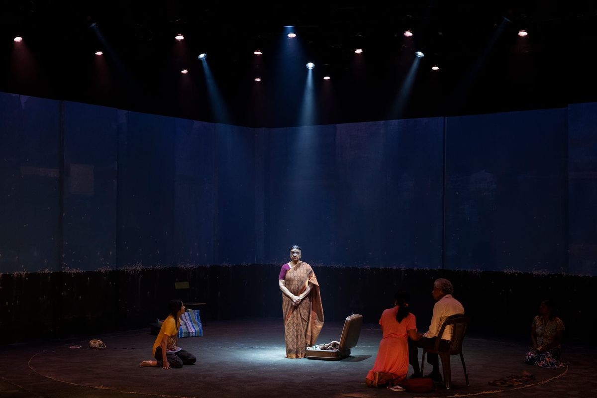 Gowrie stands alone on the wide, dimly lit stage, surrounded by darkness and the curved industrial set.