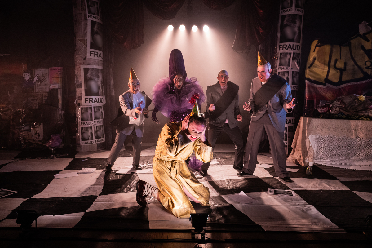 Five performers in party hats and exaggerated costumes crowd around a noseless figure crouched on the floor with hands over ears, amid a ramshackle set plastered with ‘FRAUD’ posters.