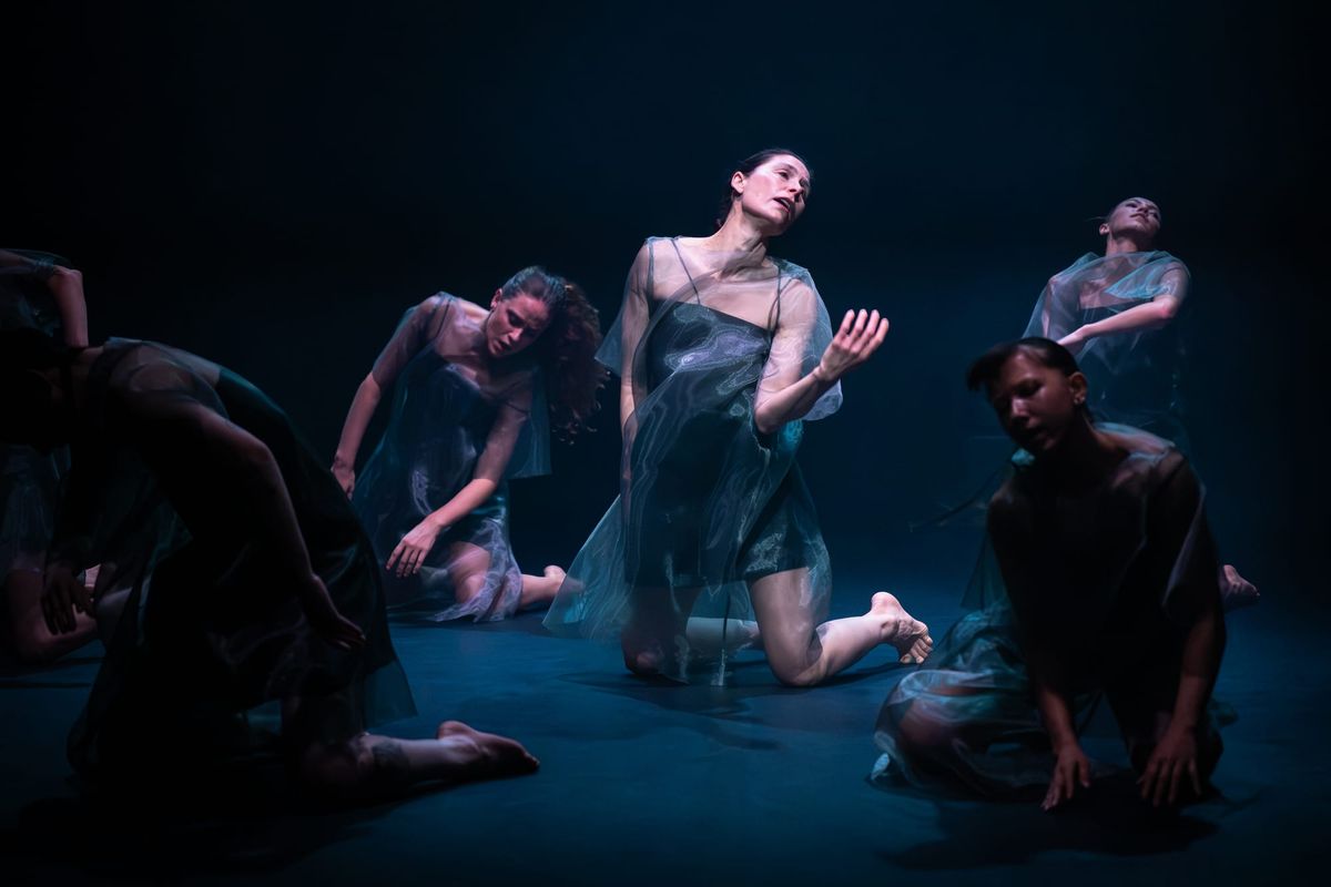 Deanne Butterworth kneels at centre among six dancers, their bodies gathered in different poses under cool blue stage lighting in 7 Dances for 7 People.