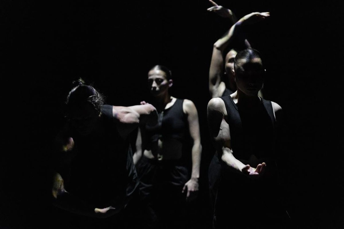 Four dancers from Triptych emerge from near-darkness, their black costumes and partially lit bodies creating fragmented shapes against the black stage.