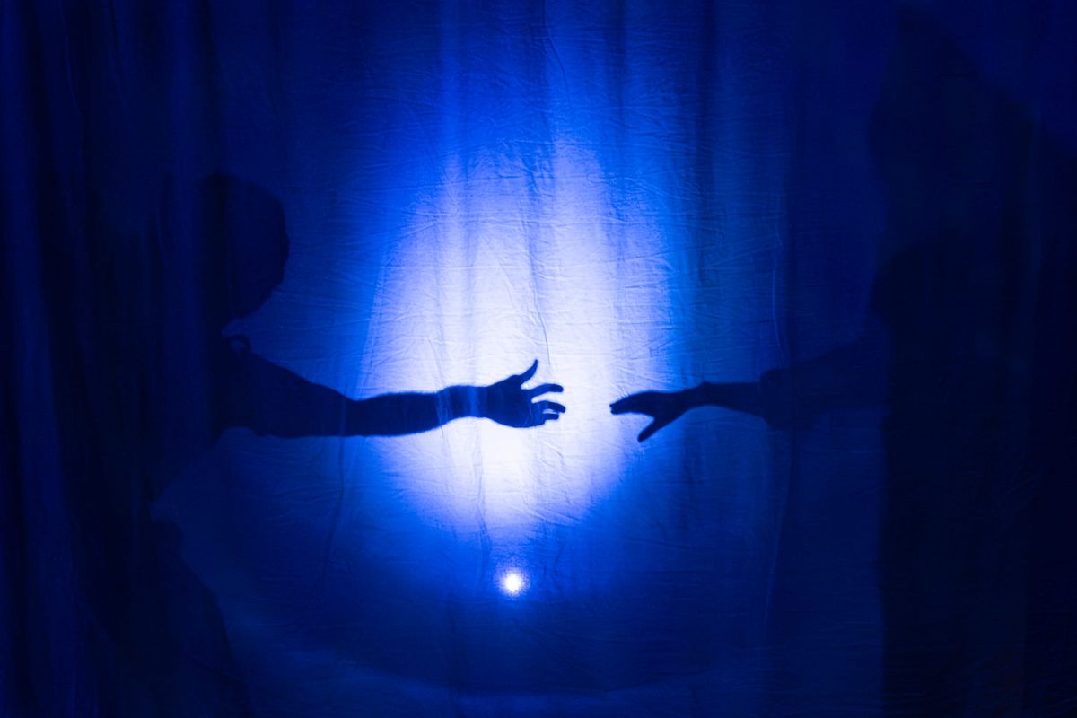 Two silhouetted figures reach towards one another behind a translucent screen washed in deep blue light during The Memory Mirror.