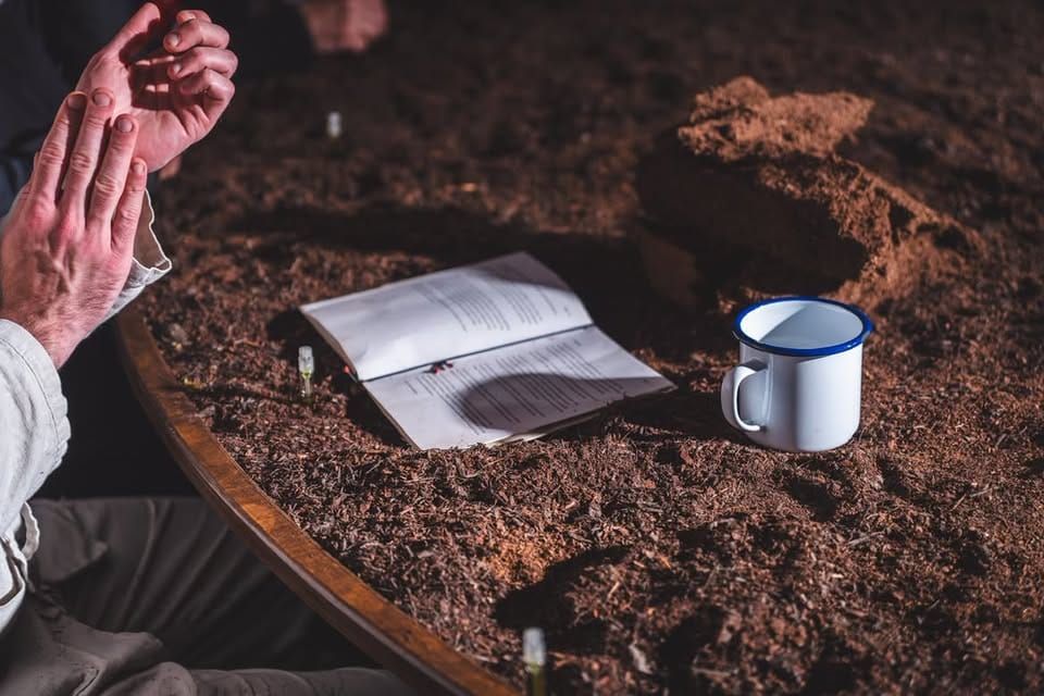 Luke Casserly sits beside a large circular table covered in dark bog soil, his hands raised above an open book and enamel mug.