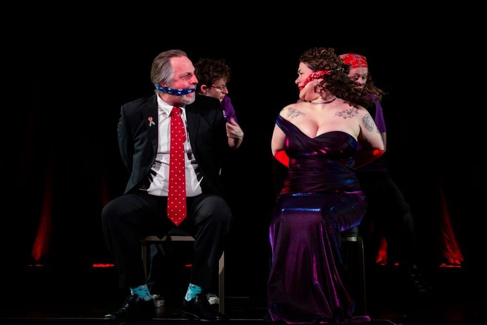 Scott Gooding as Richard Head and Mon Wing-Yun as Philanthropy Philharmonica sit bound and gagged onstage, with two ACT members standing behind them.