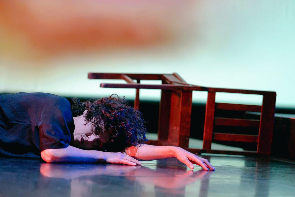 Anna Seymour lies curled on the stage floor, one arm stretched out towards the camera, with an overturned wooden chair behind her. Soft red and blue light falls across the scene.