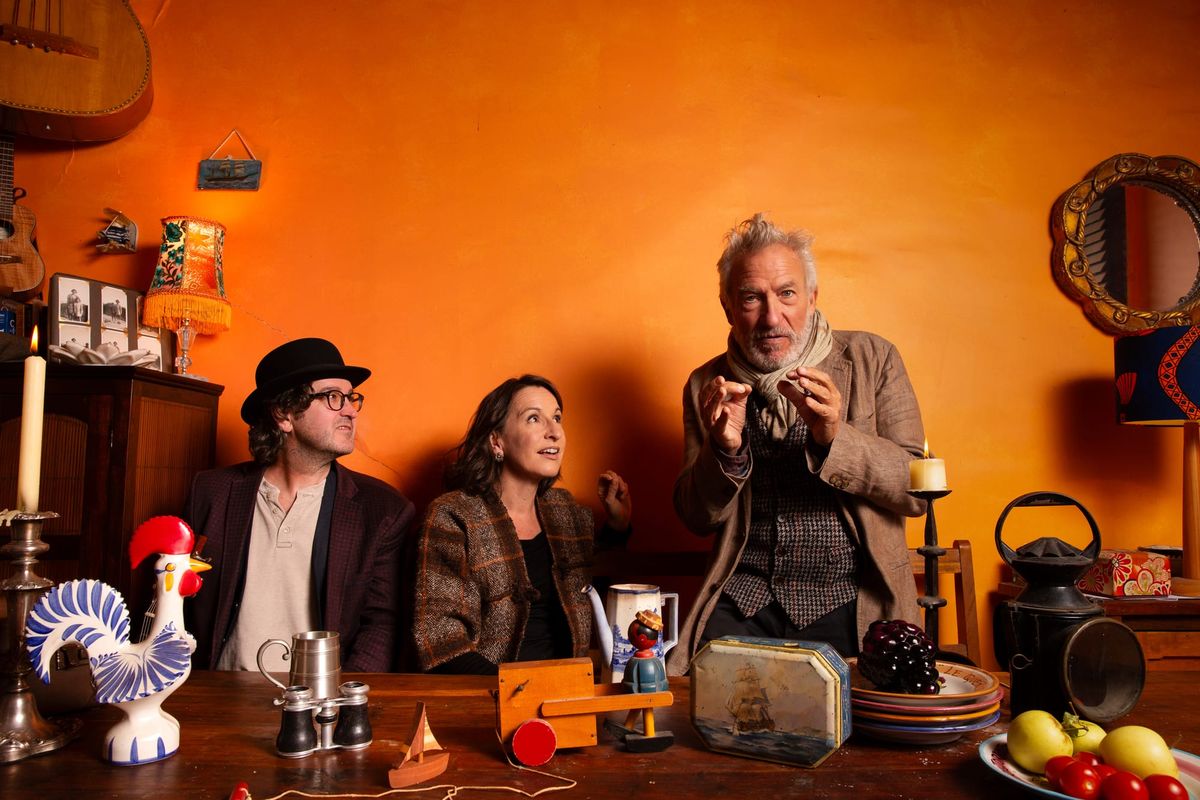 Robbie Melville and Zoe Knighton watch Richard Piper gesture animatedly behind a table crowded with wooden toys, crockery, fruit and a ceramic rooster, against a vivid orange wall.