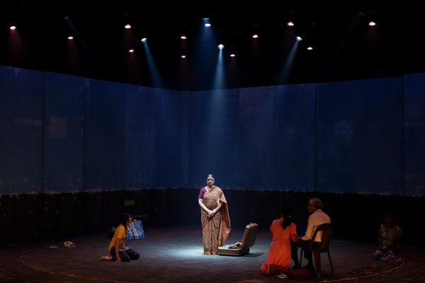 Gowrie stands alone on the wide, dimly lit stage, surrounded by darkness and the curved industrial set.