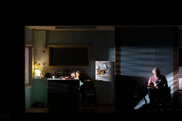 Rae sleeps on the kitchen bench while Pop sleeps nearby in his chair inside Pamela’s dimly lit home, surrounded by the darkness of the stage.