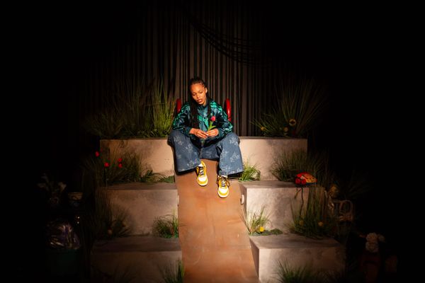 Lucy Ansell sits alone on a raised concrete walkway surrounded by flowers and plants in Red Stitch's production of Funeral Flowers.