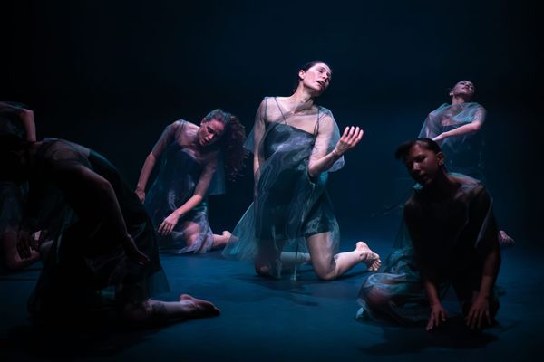 Deanne Butterworth kneels at centre among six dancers, their bodies gathered in different poses under cool blue stage lighting in 7 Dances for 7 People.