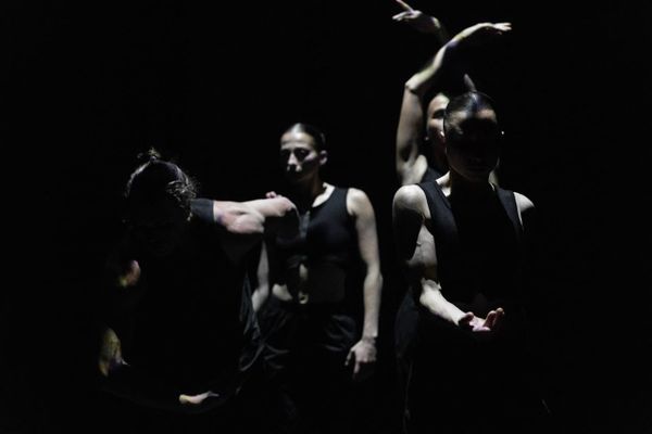 Four dancers from Triptych emerge from near-darkness, their black costumes and partially lit bodies creating fragmented shapes against the black stage.