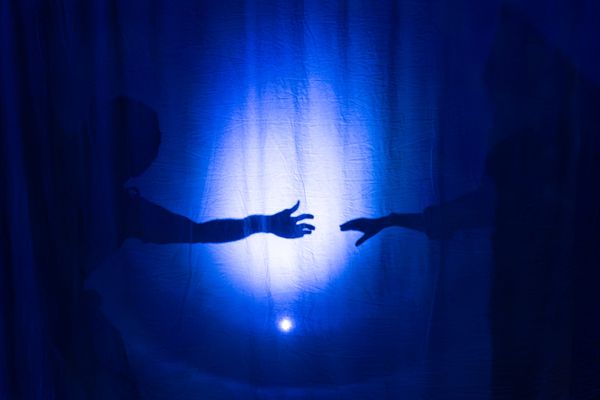 Two silhouetted figures reach towards one another behind a translucent screen washed in deep blue light during The Memory Mirror.