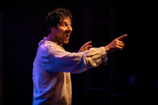 Fabio Motta in Holy Fool, leaning forward with an exuberant expression and pointing across the darkened stage.