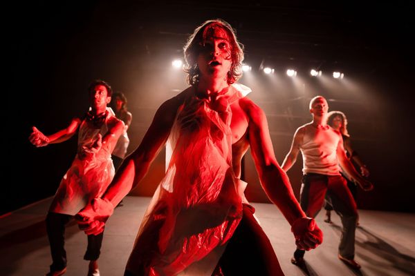 Three performers close up under intense red lighting, with other members of the Lord of the Flies ensemble behind them on the raked stage.