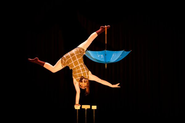 Holly Muir balances upside down on handstand canes, one leg extended beneath a blue umbrella.